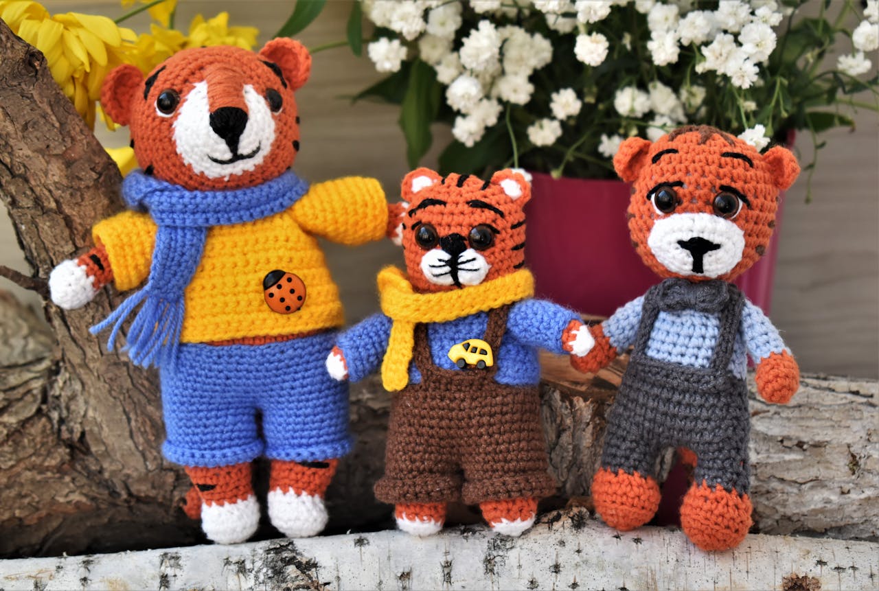 Three knitted teddy bear toys in vibrant clothing, posed on a wooden log with flowers in the background.